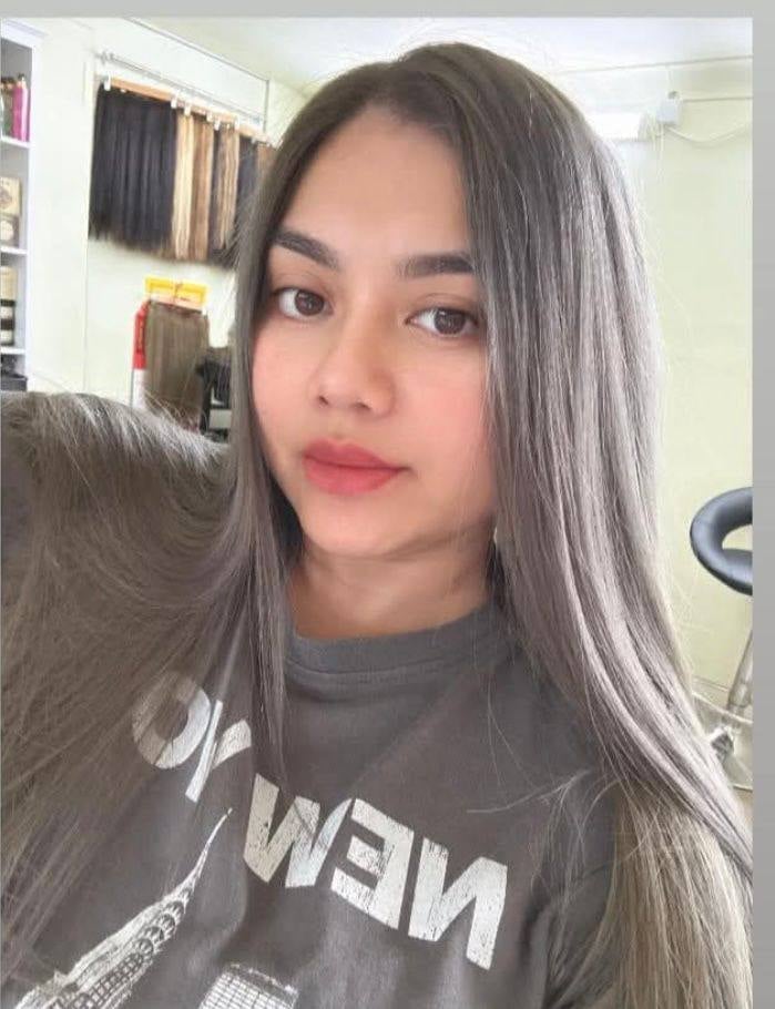 Young woman with long dark hair wearing a dark graphic t-shirt, taking a selfie indoors with shelving in the background