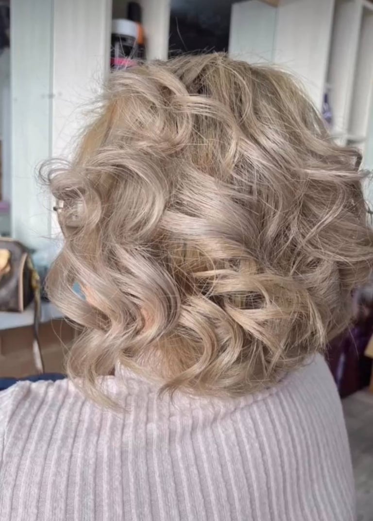 Back view of a woman with long, wavy blonde hair styled in loose curls, wearing a light purple ribbed sweater