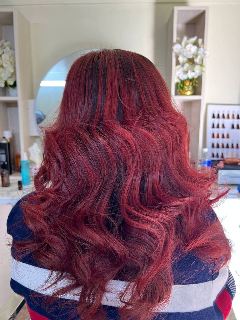 Back view of person with long burgundy wavy hair in a salon setting with shelves of nail polish and products