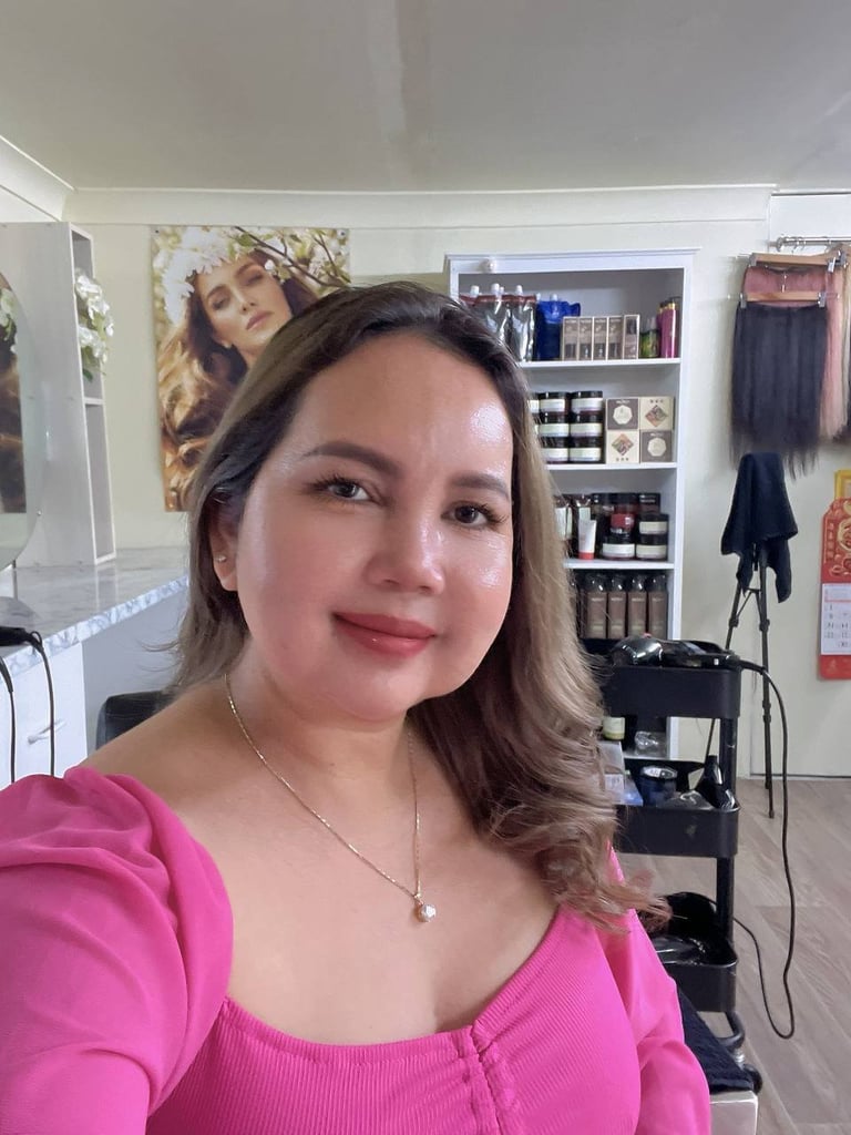 Woman in bright pink top taking a selfie in a beauty room with shelves of makeup, hair tools, and styling products in background