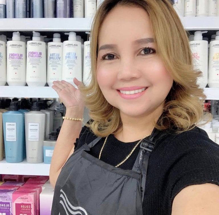 Woman with blonde wavy hair smiling at camera wearing black apron, standing in front of shelves of beauty and hair care products