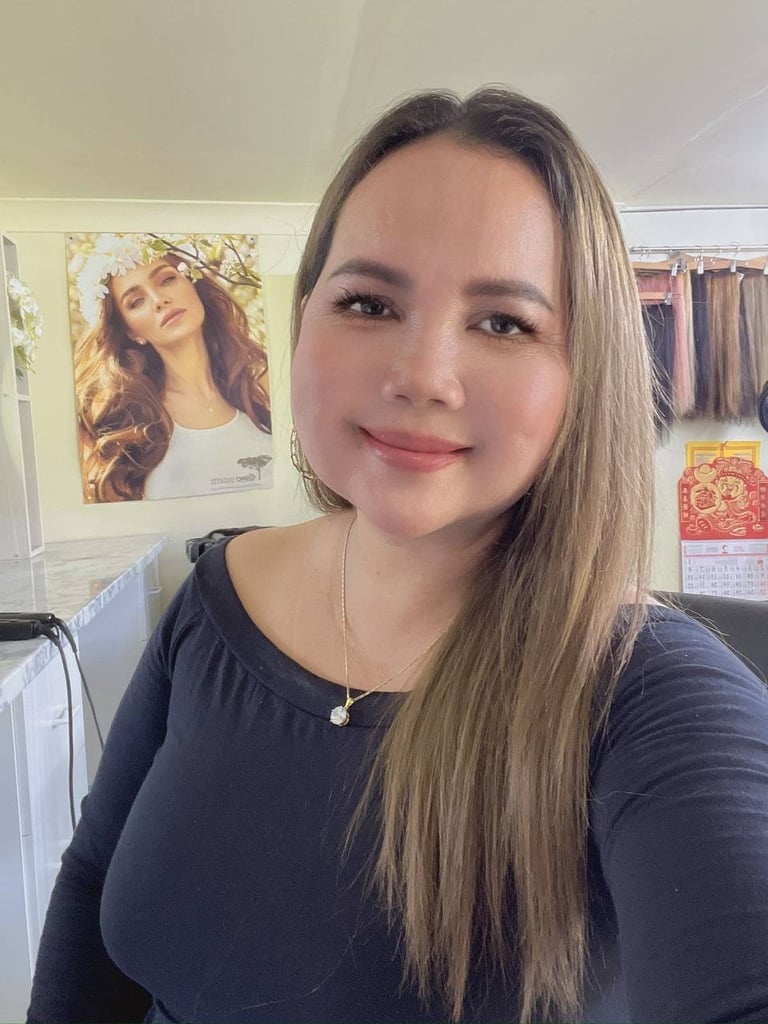 Young woman with long brown hair wearing a dark top and pendant necklace, smiling at camera in an indoor room setting